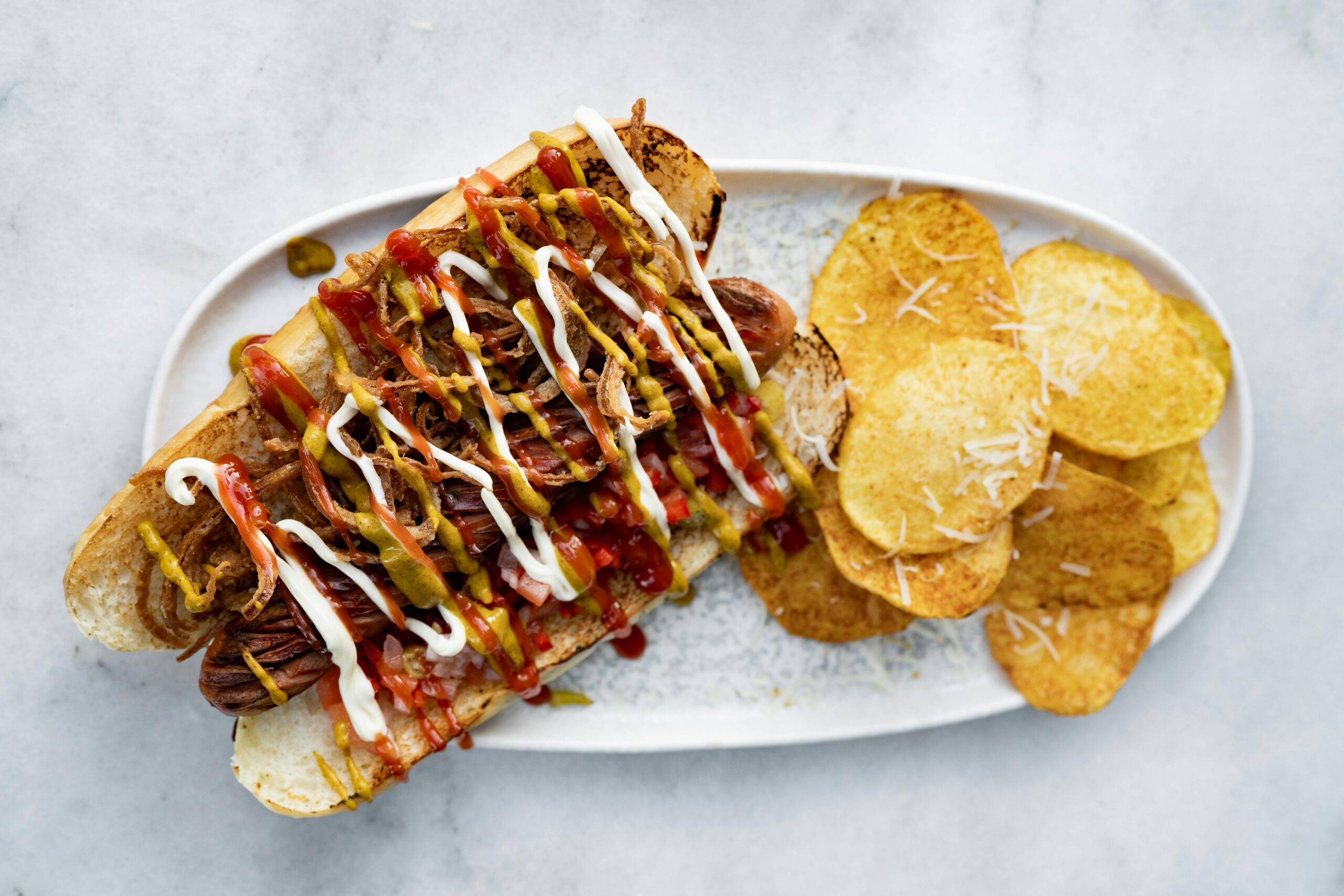 Hotdog with different sauces drizzled on top and a side of chips.